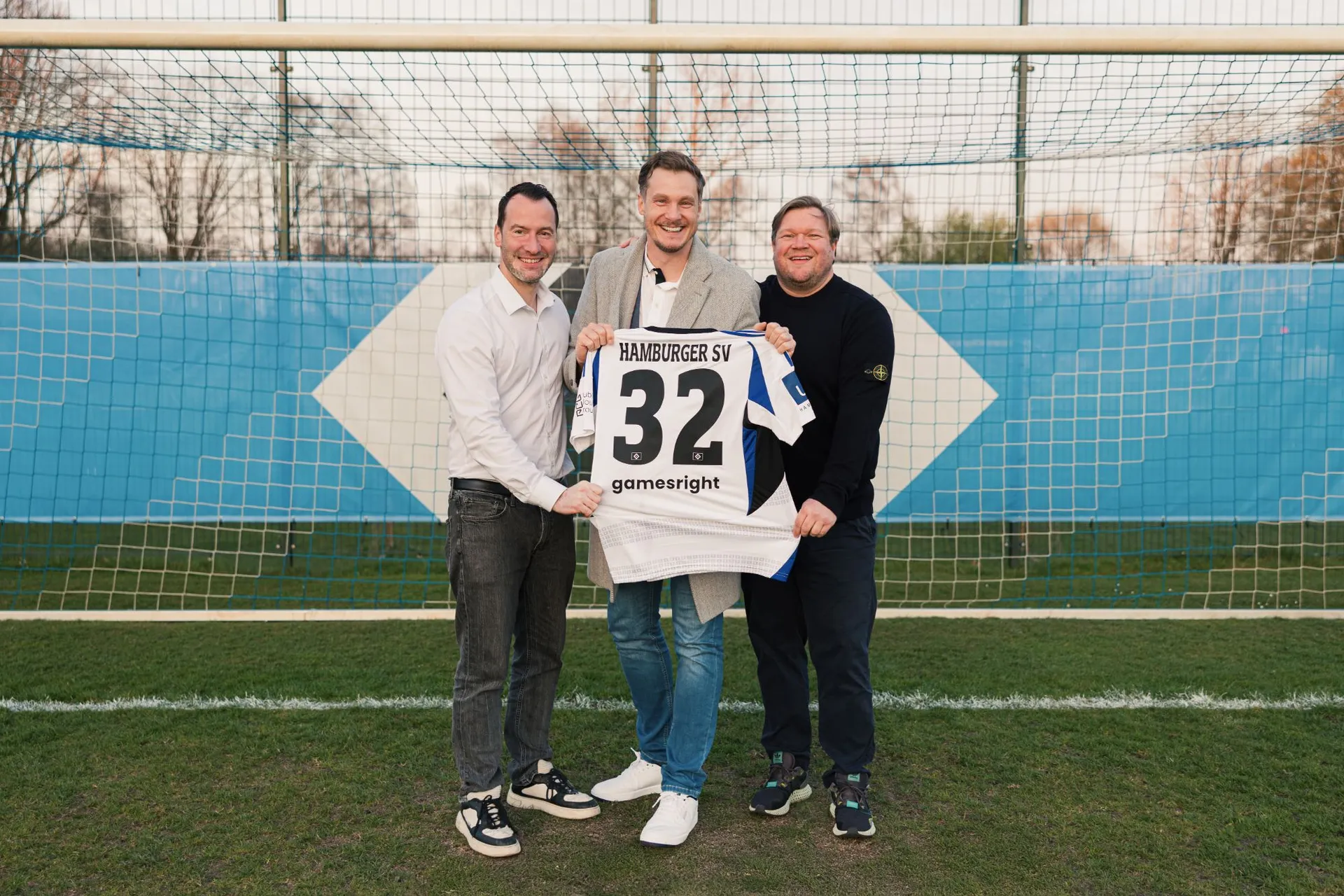 Drei Männer halten lachend ein HSV-Trikot mit der Nummer 32 und dem gamesright-Schriftzug vor einem Tor.