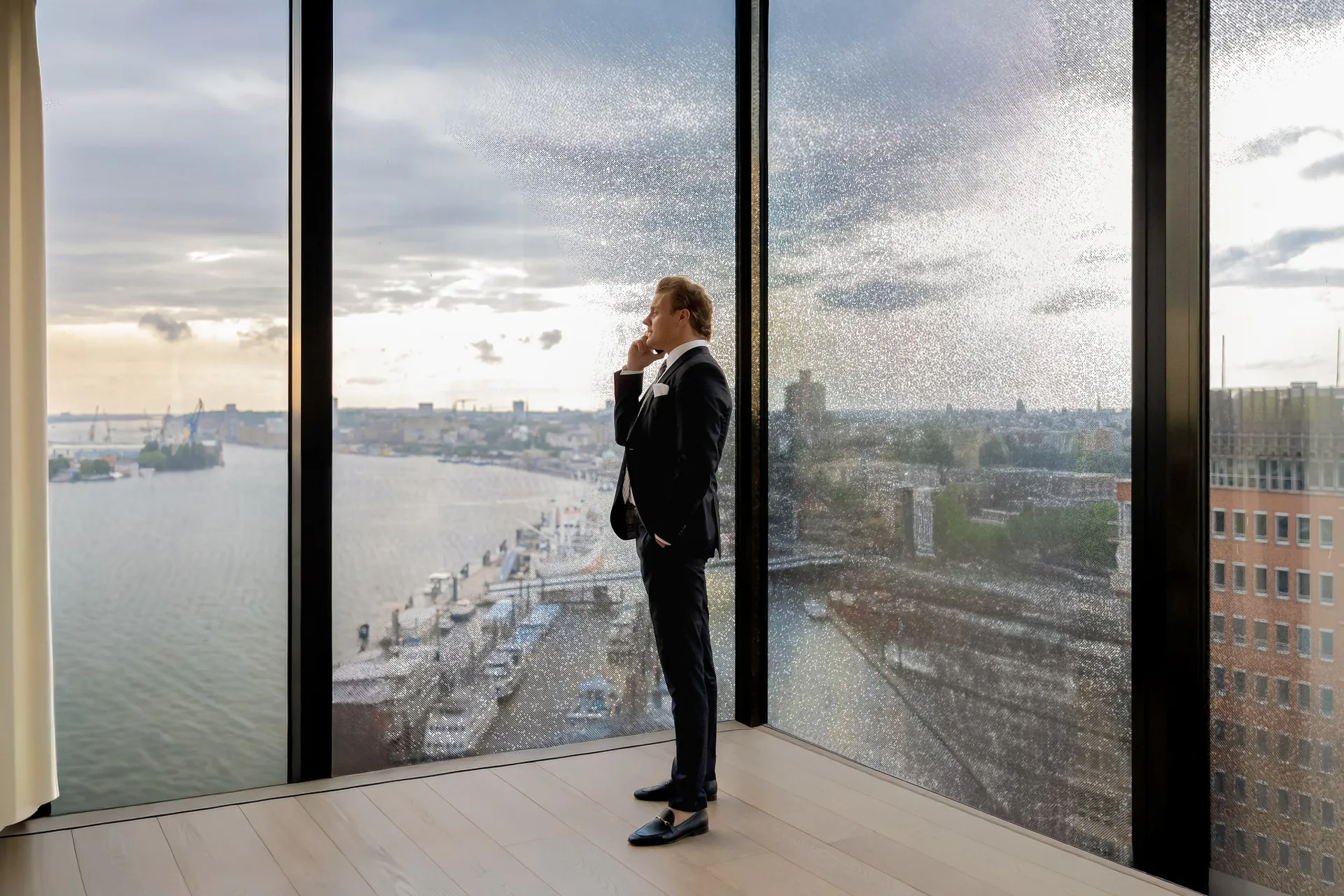 Geschäftsmann im Anzug telefoniert an einer bodentiefen Fensterfront hoch über dem Hamburger Hafen.